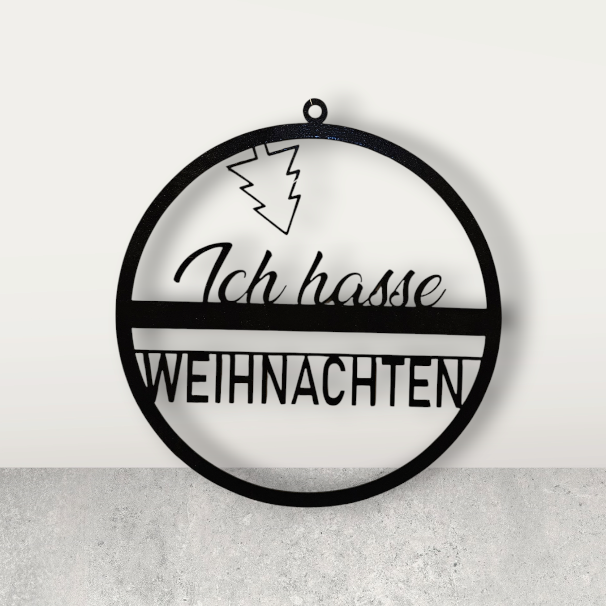 Eine kreisförmige Türdekoration, in der Mitte der Text "Ich hasse Weihnachten" in 2 verschiedenen Schriftarten, oben ist ein Weihnachtsbaum kopüber angebracht". Schwarz